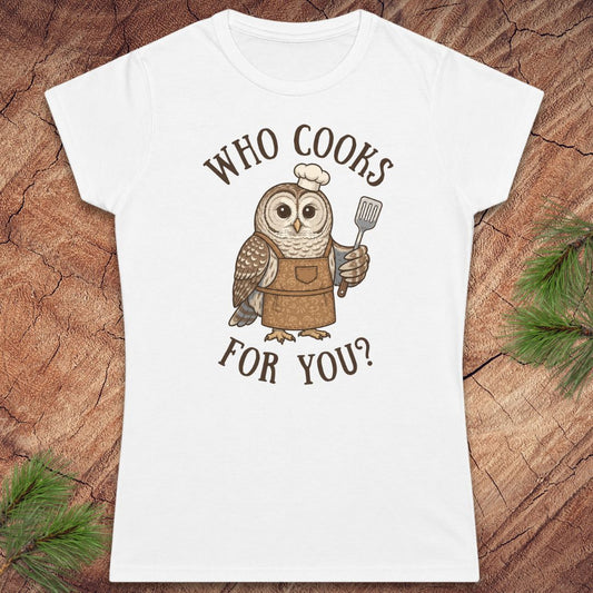 A white short-sleeve crewneck t-shirt featuring a cartoon owl wearing a brown apron and chef's hat, holding a spatula, with the text 'WHO COOKS FOR YOU?' printed in brown. The shirt is displayed flat on a textured wood background with pine needles in the corners.