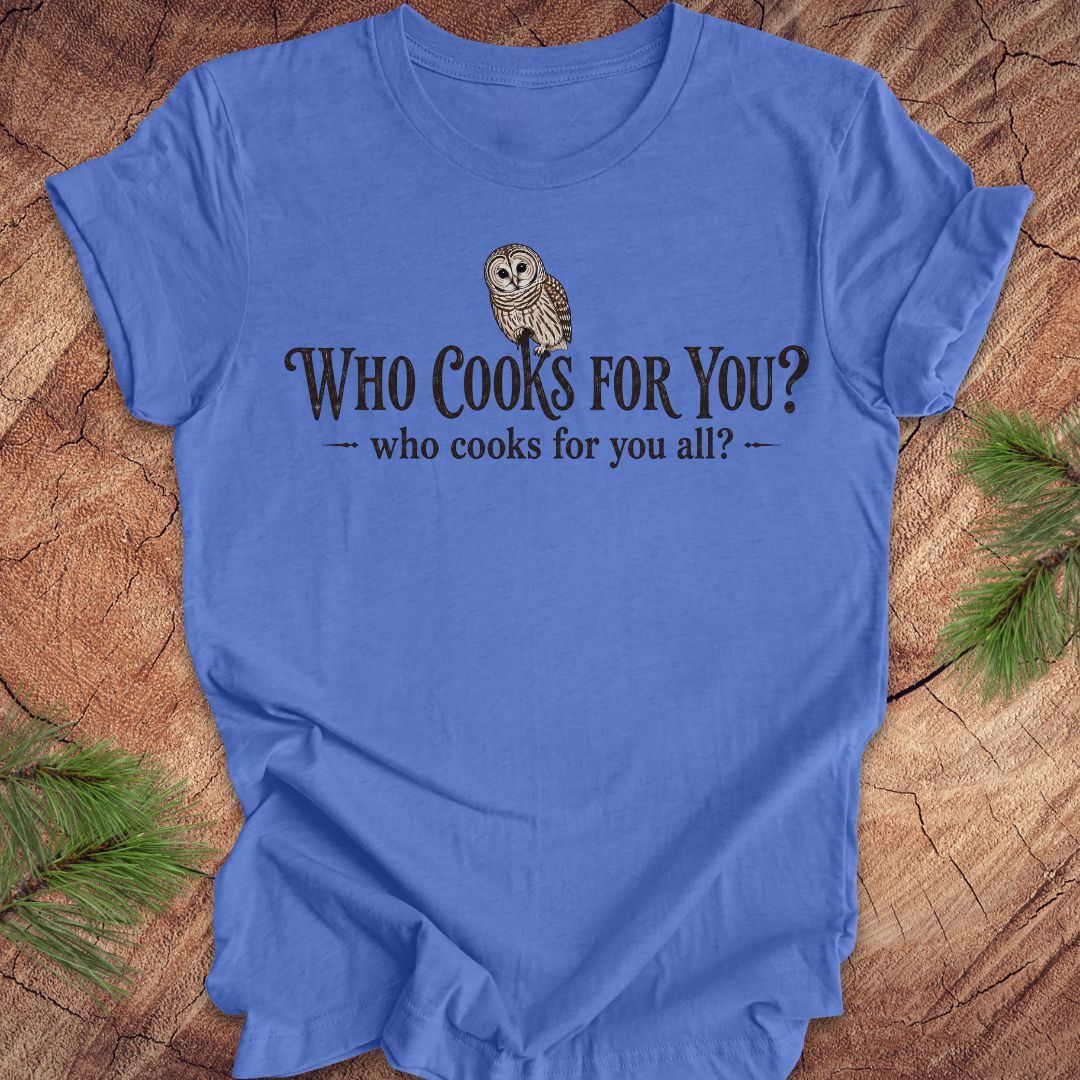 Blue t-shirt with an owl and text, 'who cooks for you?' on a wood background.