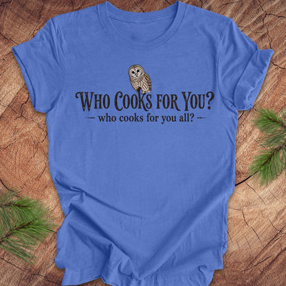 Blue t-shirt with an owl and text, 'who cooks for you?' on a wood background.