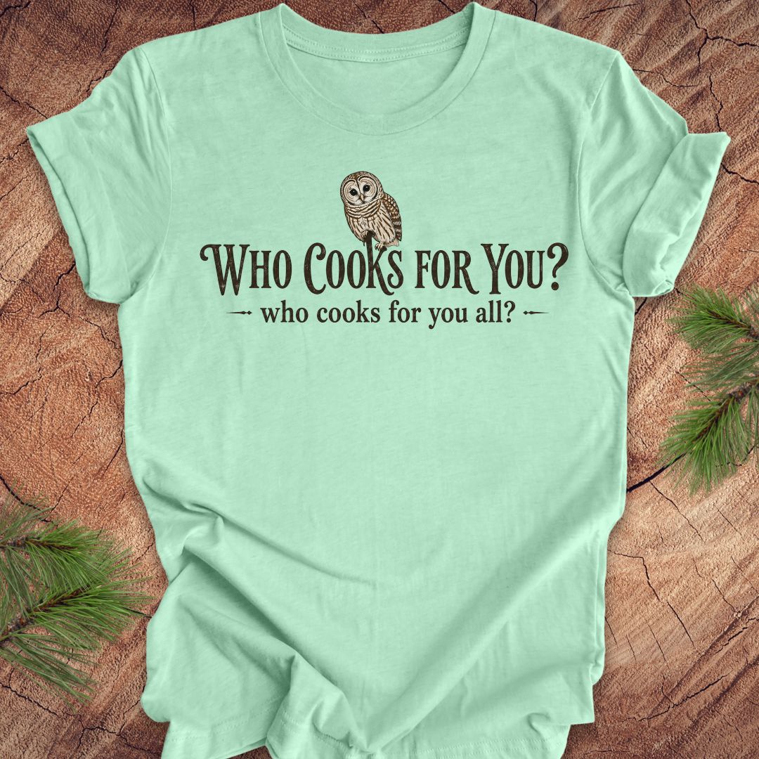 Mint green t-shirt with an owl and text, 'who cooks for you?' on a wood background.