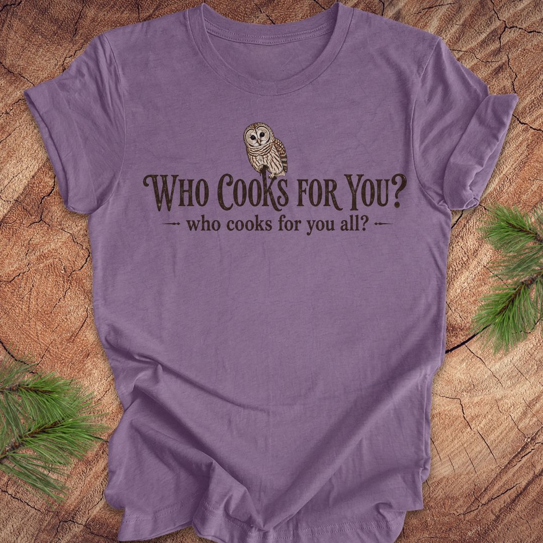 Purple t-shirt with an owl and text, 'who cooks for you?' on a wood background.