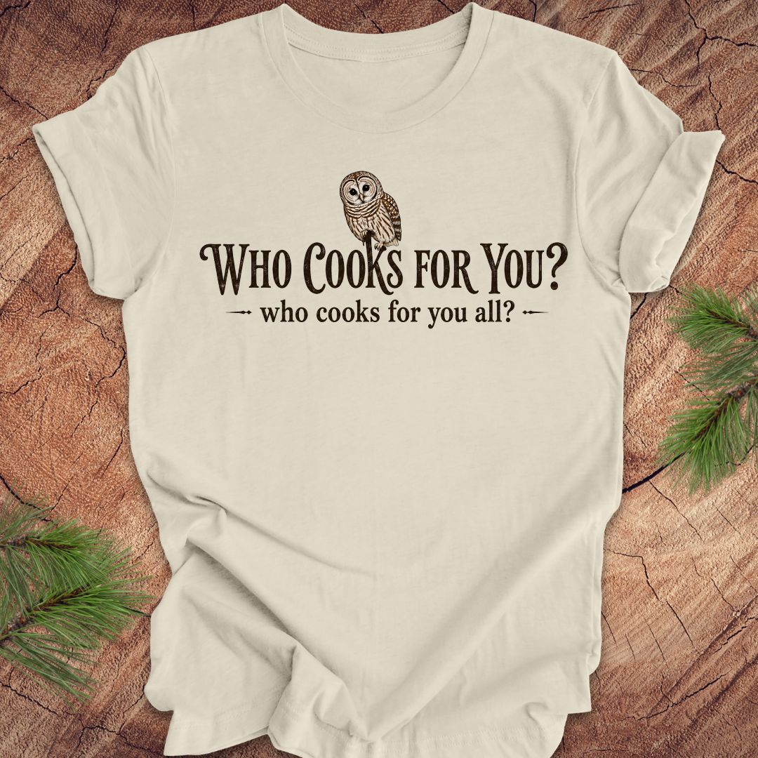 Beige t-shirt with an owl and text, 'who cooks for you?' on a wood background.