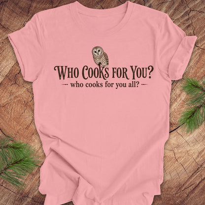 Pink t-shirt with an owl and text, 'who cooks for you?' on a wood background.