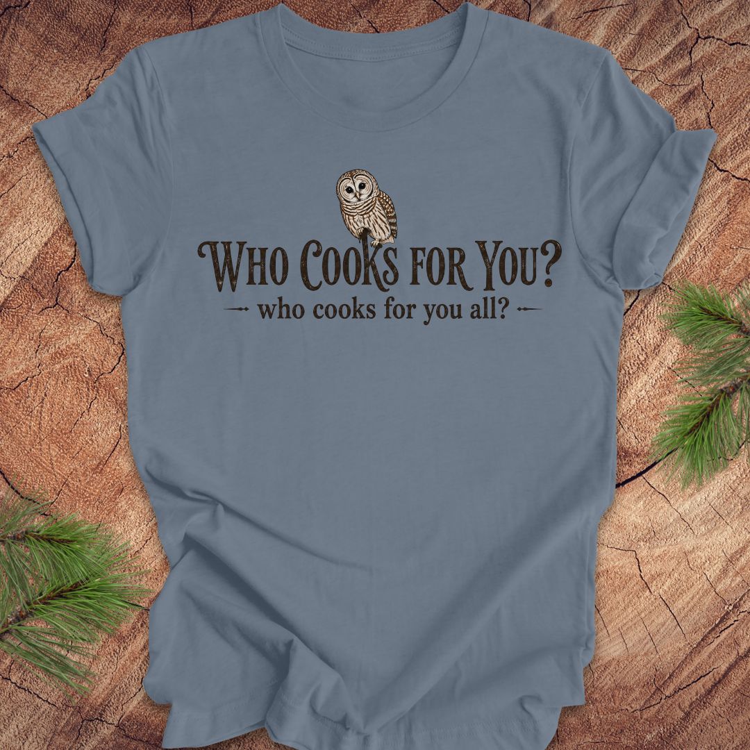 Blue t-shirt with an owl and text, 'who cooks for you?' on a wood background.