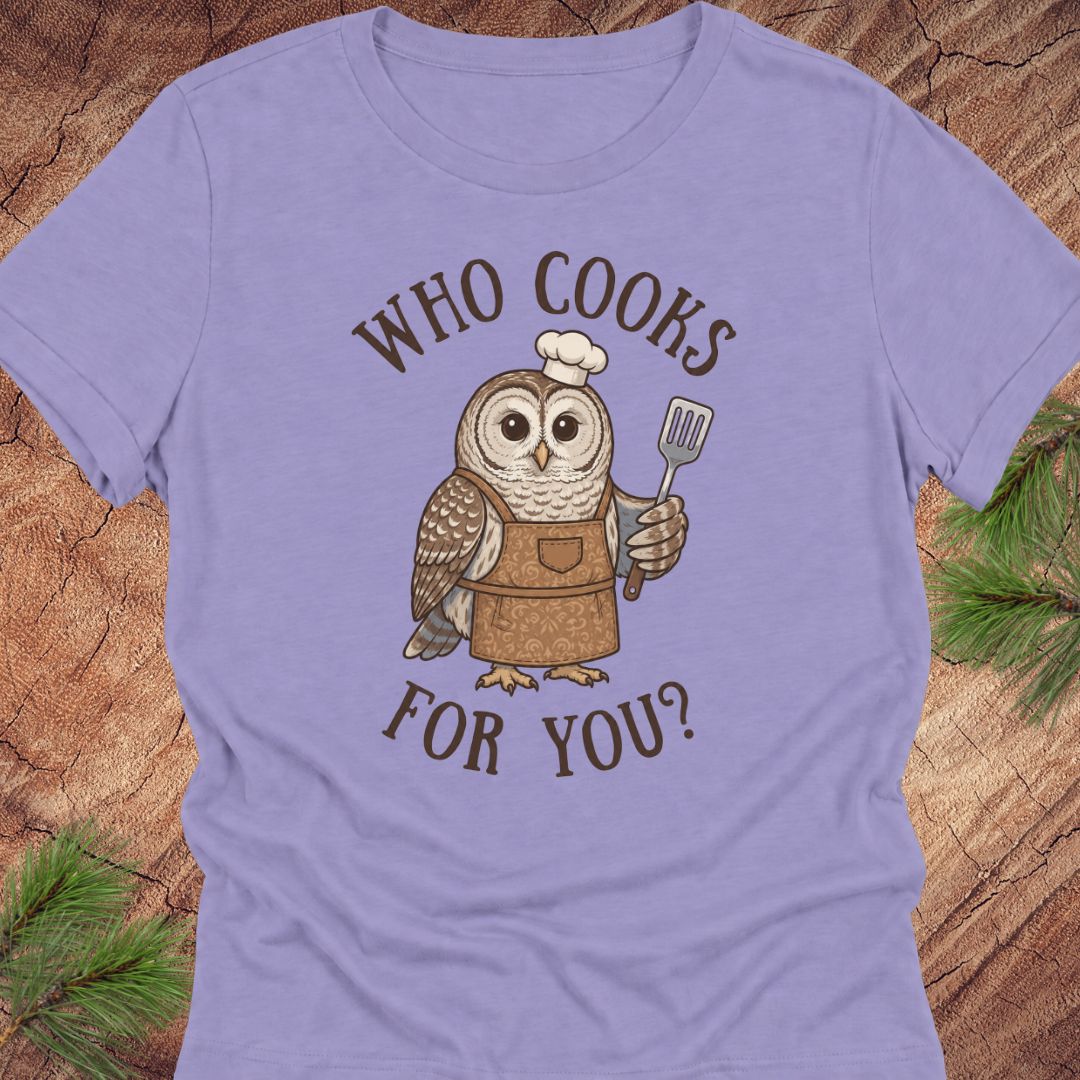 A lavender short-sleeve crewneck t-shirt featuring a cartoon owl wearing a brown apron and white chef's hat, holding a spatula, with the text 'WHO COOKS FOR YOU?' in brown. The shirt is laid flat on a rustic wood background with pine needles in the corners.