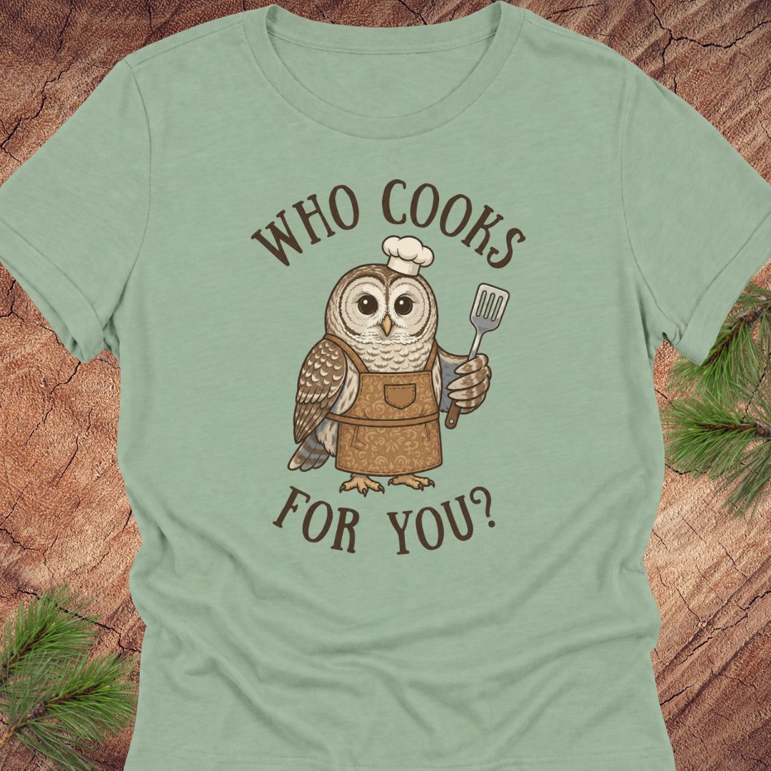 A sage green short-sleeve t-shirt with a graphic of an owl wearing a chef's hat and apron, holding a spatula, with the text 'WHO COOKS FOR YOU?' in brown. The shirt is displayed flat on a rustic wood background with pine needles in the corners.