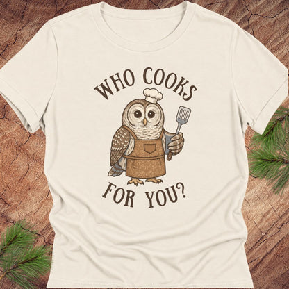An off-white short-sleeve t-shirt with a graphic of a cute owl wearing a brown apron and white chef's hat, holding a spatula. The text 'WHO COOKS FOR YOU?' is printed in brown around the owl.