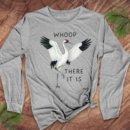 A heather gray long-sleeve t-shirt with a graphic of a whooping crane and the text 'WHOOP THERE IT IS', laid flat on a rustic wood background with pine needles.