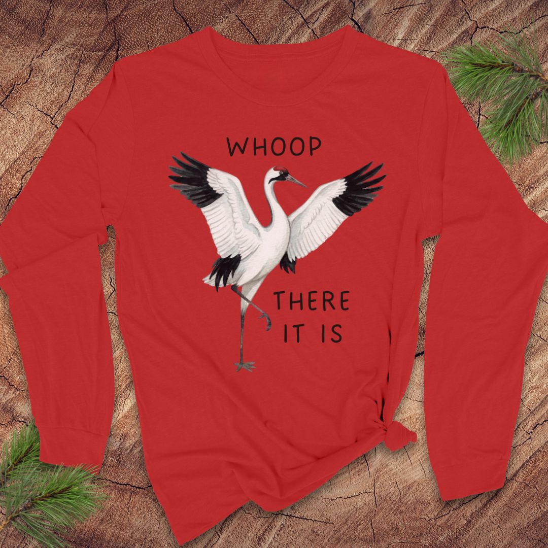 A red long-sleeve crewneck t-shirt featuring a graphic of a whooping crane with black-tipped wings and the text 'WHOOP THERE IT IS' in black. The t-shirt is styled flat on a rustic wood background with pine needles in the corners.