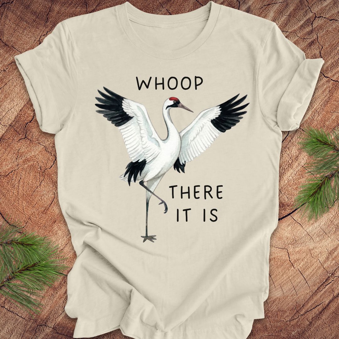 Beige t-shirt with a crane graphic and text, 'whoop there it is' on a wooden surface