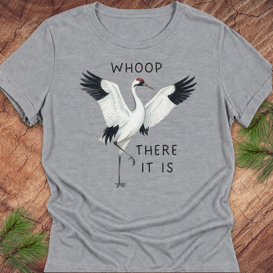 A heather gray short-sleeve t-shirt featuring a graphic of a red-crowned crane with outstretched wings and the text 'WHOOP THERE IT IS' printed on the front. The t-shirt is laid flat on a rustic wood background with pine needles in the corners.