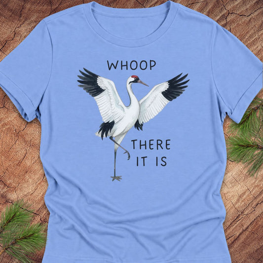 A light blue short-sleeve crewneck t-shirt featuring a graphic of a white crane with black wings and the text 'WHOOP THERE IT IS' printed on the front, laid flat on a rustic wood background with pine needles in the corners.