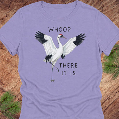 A lavender purple short-sleeve t-shirt featuring a graphic of two cranes with the text 'WHOOP THERE IT IS' printed on the front, laid flat on a rustic wood background with pine needles.