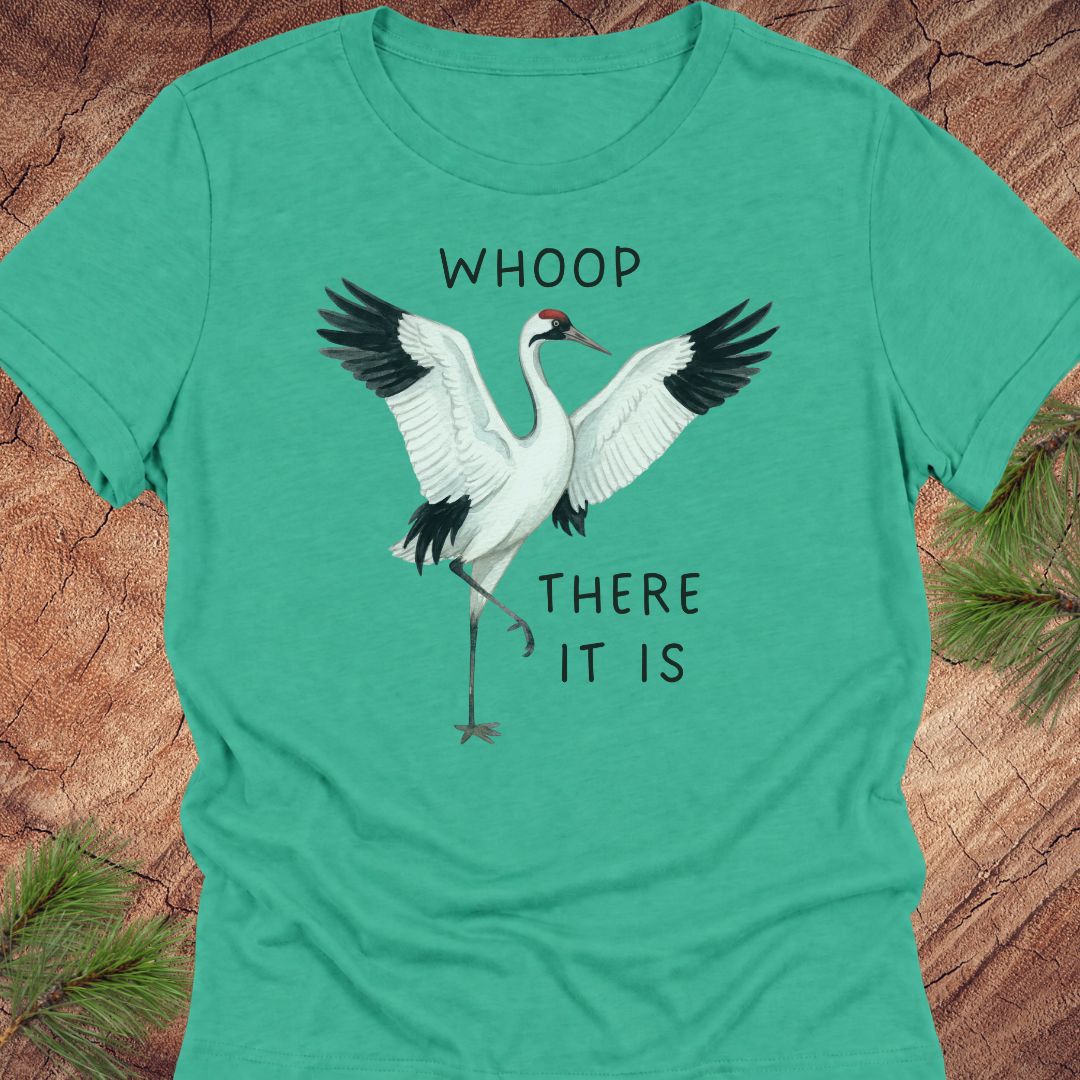 A green short-sleeve crewneck t-shirt featuring a graphic of a white crane with black wingtips and the text 'WHOOP THERE IT IS' printed on the front, laid flat on a rustic wood background with pine needles.