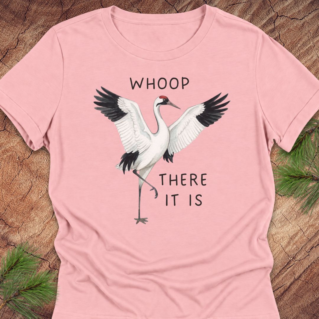 A light pink short-sleeve t-shirt with a graphic of a white crane with black-tipped wings and the text 'WHOOP THERE IT IS' printed on the front, laid flat on a rustic wood background with pine needles in the corners.