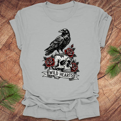 Silver t-shirt with a crow, skull, and roses design with text, 'wild hearts' on a wooden surface.