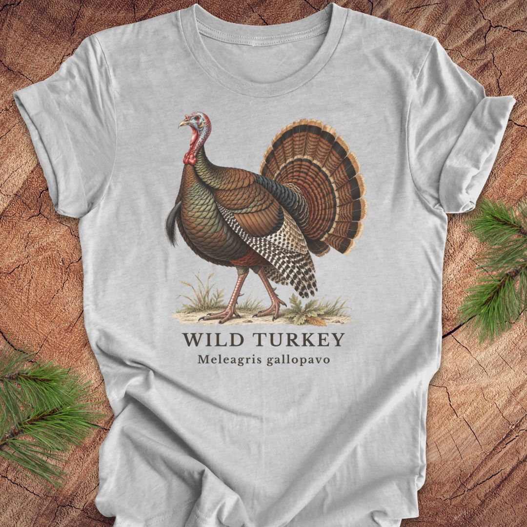A light gray short-sleeve t-shirt with a vintage-style illustration of a wild turkey and the text 'WILD TURKEY' and 'Meleagris gallopavo' printed on the front, displayed flat on a rustic wood background with pine branches in the corners.