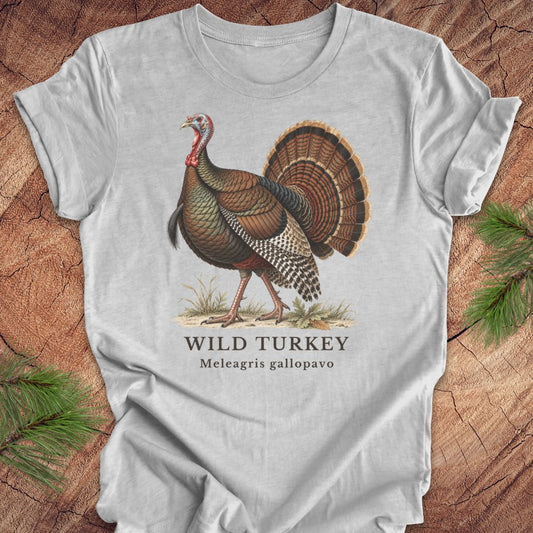 A light gray short-sleeve t-shirt with a vintage-style illustration of a wild turkey and the text 'WILD TURKEY' and 'Meleagris gallopavo' printed on the front, displayed flat on a rustic wood background with pine branches in the corners.