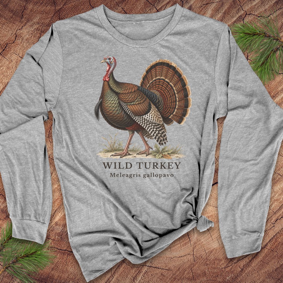 Athletic Heather gray long-sleeve t-shirt with a vintage naturalist-style wild turkey illustration labeled 'Wild Turkey, Meleagris gallopavo', laid flat on a rustic wood background with pine branches.