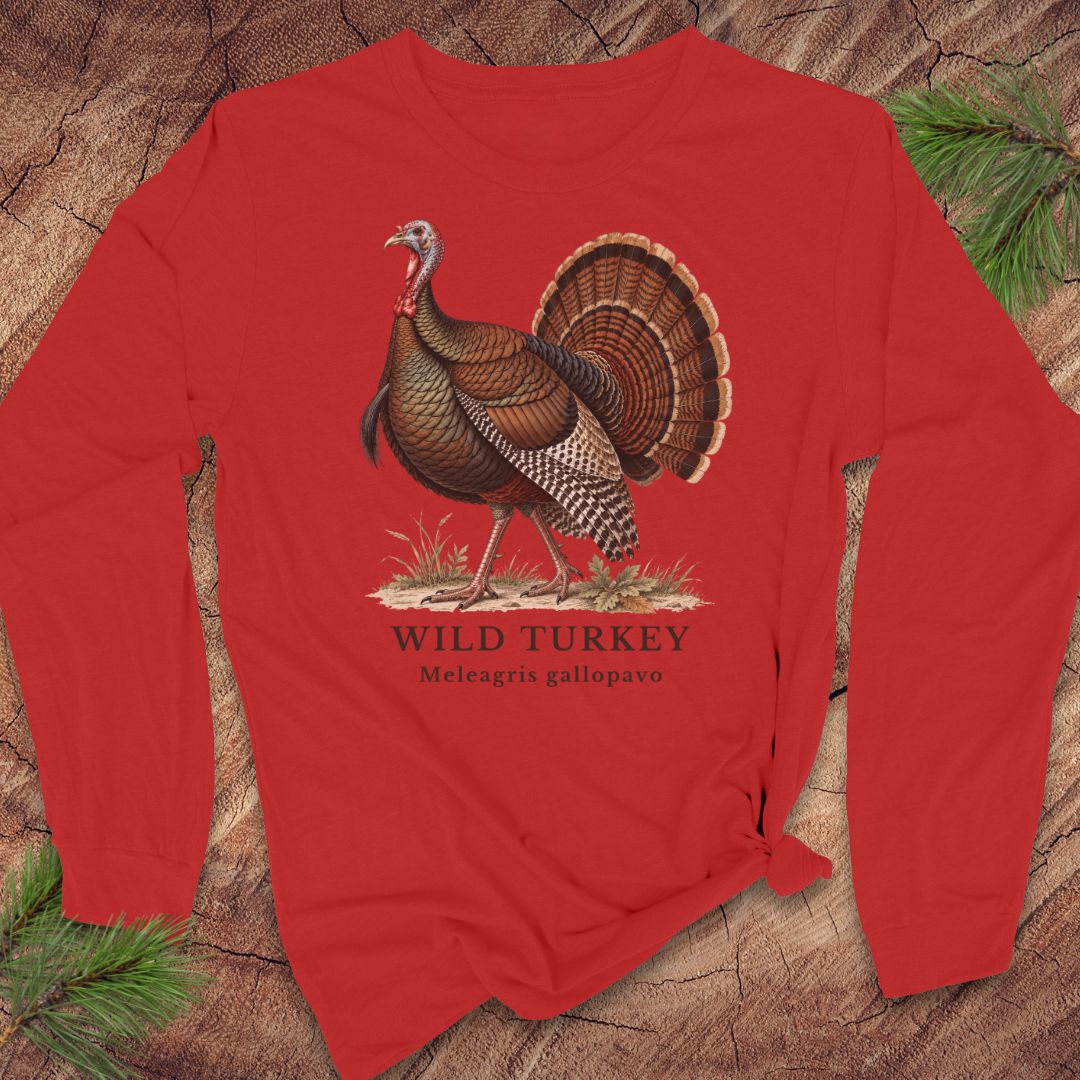 Red long-sleeve t-shirt with a vintage naturalist-style wild turkey illustration labeled 'Wild Turkey, Meleagris gallopavo', laid flat on a rustic wood background with pine branches.