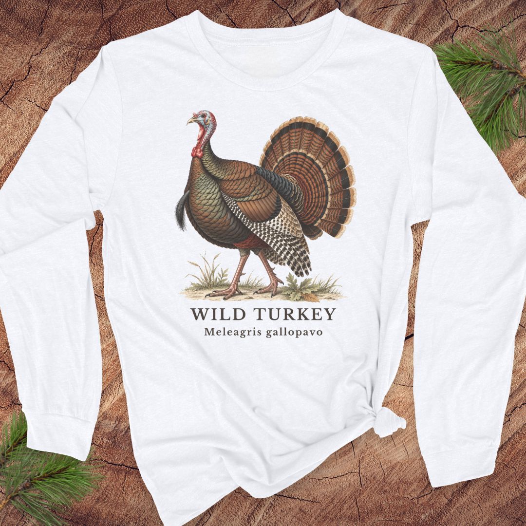 White long-sleeve t-shirt with a vintage naturalist-style wild turkey illustration labeled 'Wild Turkey, Meleagris gallopavo', laid flat on a rustic wood background with pine branches.