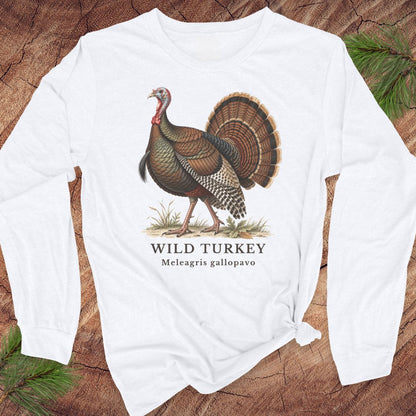 White long-sleeve t-shirt with a vintage naturalist-style wild turkey illustration labeled 'Wild Turkey, Meleagris gallopavo', laid flat on a rustic wood background with pine branches.