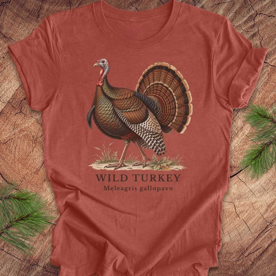 A rust-colored short-sleeve t-shirt with a vintage-style illustration of a wild turkey on the front, laid flat on a rustic wood background with pine branches in the corners.