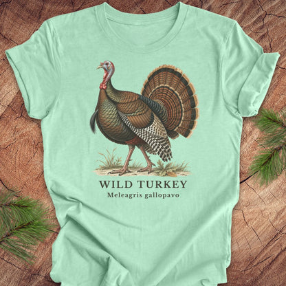 A mint green short-sleeve t-shirt with a vintage-style illustration of a wild turkey and the text 'WILD TURKEY' and 'Meleagris gallopavo' printed on the front, laid flat on a rustic wood background with pine branches in the corners.