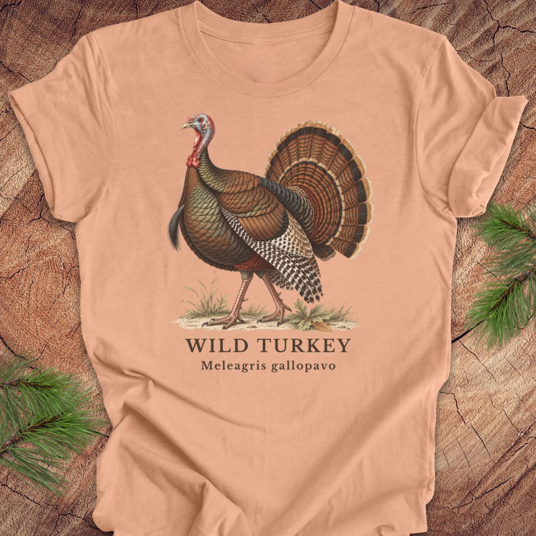 A peach-colored short-sleeve t-shirt laid flat on a rustic wood background with pine branches in the corners. The shirt features a vintage-style illustration of a wild turkey with the text 'WILD TURKEY' and 'Meleagris gallopavo' printed below.