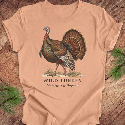 A peach-colored short-sleeve t-shirt laid flat on a rustic wood background with pine branches in the corners. The shirt features a vintage-style illustration of a wild turkey with the text 'WILD TURKEY' and 'Meleagris gallopavo' printed below.