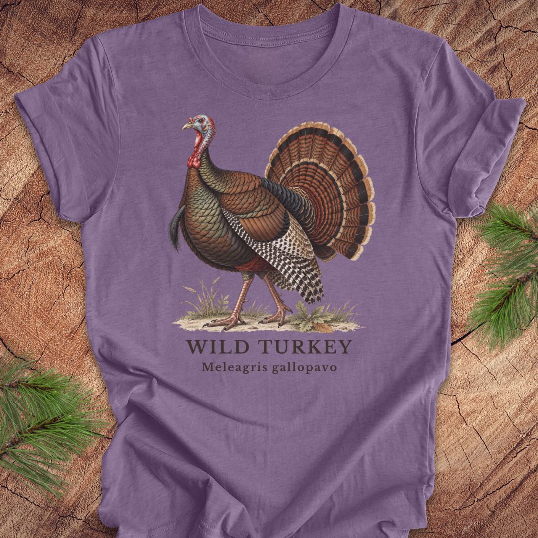 A purple short-sleeve t-shirt laid flat on a rustic wood background with pine branches at the corners. The shirt features a vintage-style illustration of a wild turkey with the text 'WILD TURKEY' and 'Meleagris gallopavo' printed below.