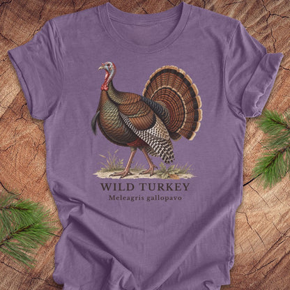 A purple short-sleeve t-shirt laid flat on a rustic wood background with pine branches at the corners. The shirt features a vintage-style illustration of a wild turkey with the text 'WILD TURKEY' and 'Meleagris gallopavo' printed below.