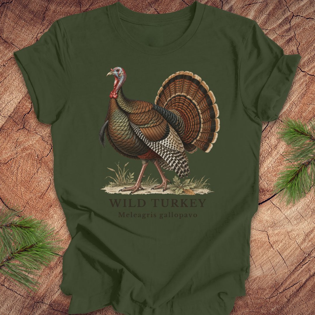 An olive green short-sleeve t-shirt featuring a detailed vintage-style graphic of a wild turkey with the text 'WILD TURKEY' and 'Meleagris gallopavo' printed below it. The t-shirt is displayed flat on a rustic wood background decorated with pine branches.