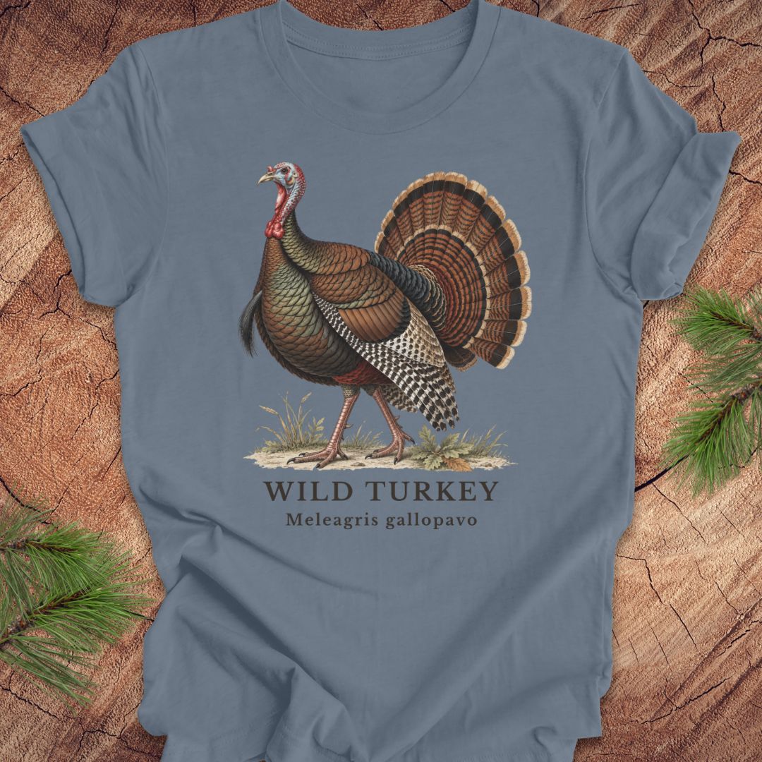 A slate blue crewneck t-shirt featuring a vintage-style illustration of a wild turkey with the text 'WILD TURKEY' and 'Meleagris gallopavo' printed below it. The t-shirt is displayed flat on a rustic wood background with pine branches in the corners.