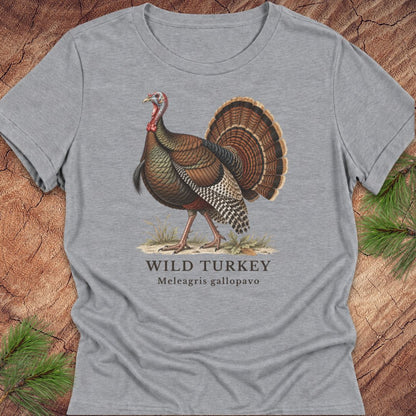 Athletic Heather gray women's relaxed t-shirt with a vintage naturalist-style wild turkey illustration labeled 'Wild Turkey, Meleagris gallopavo', laid flat on a rustic wood background with pine branches.