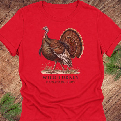 Red women's relaxed t-shirt with a vintage naturalist-style wild turkey illustration labeled 'Wild Turkey, Meleagris gallopavo', laid flat on a rustic wood background with pine branches.