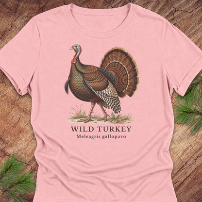 Pink women's relaxed t-shirt with a vintage naturalist-style wild turkey illustration labeled 'Wild Turkey, Meleagris gallopavo', laid flat on a rustic wood background with pine branches.
