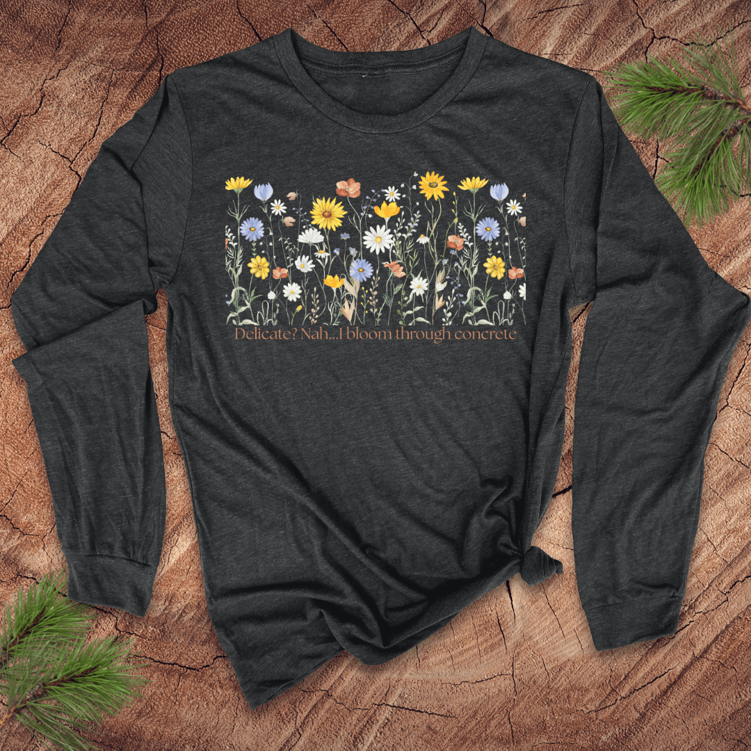 Dark gray  long sleeve tshirt with floral design and text 'delicate? nah. i bloom through concrete.' on a wooden surface.