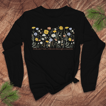 Black long sleeve tshirt with floral design and text 'delicate? nah. i bloom through concrete.' on a wooden surface.