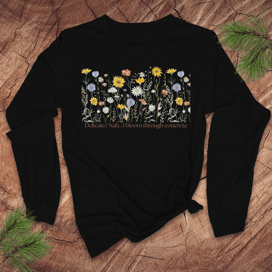 Black long sleeve tshirt with floral design and text 'delicate? nah. i bloom through concrete.' on a wooden surface.