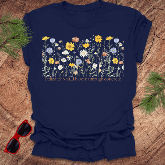 Navy t-shirt with floral design and text "delicate? nah. i bloom through concrete.' on a wooden surface.