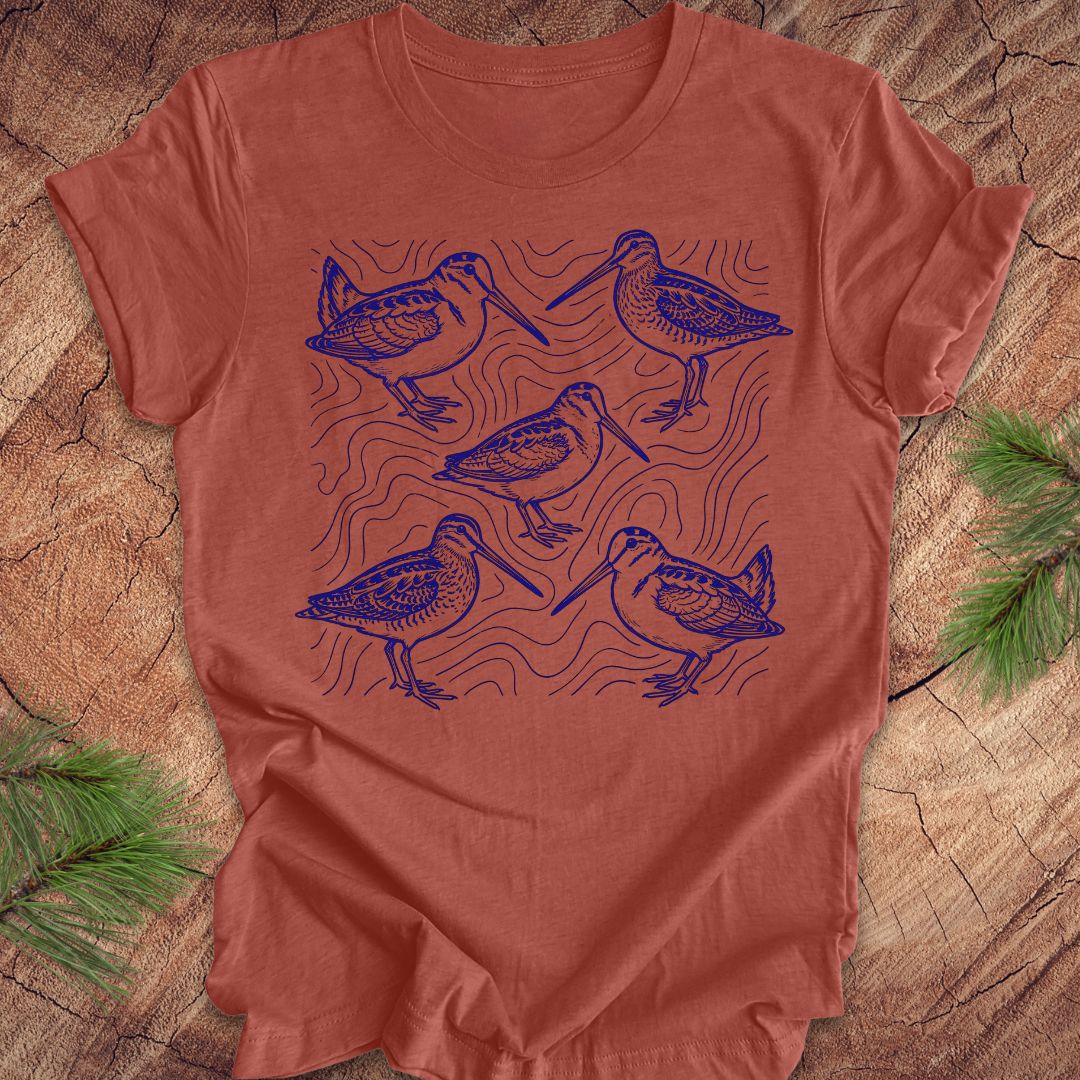 Clay colored t-shirt with blue woodcock and wilson's snipe bird illustrations on a wooden surface