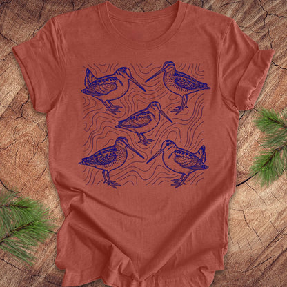 Clay colored t-shirt with blue woodcock and wilson's snipe bird illustrations on a wooden surface