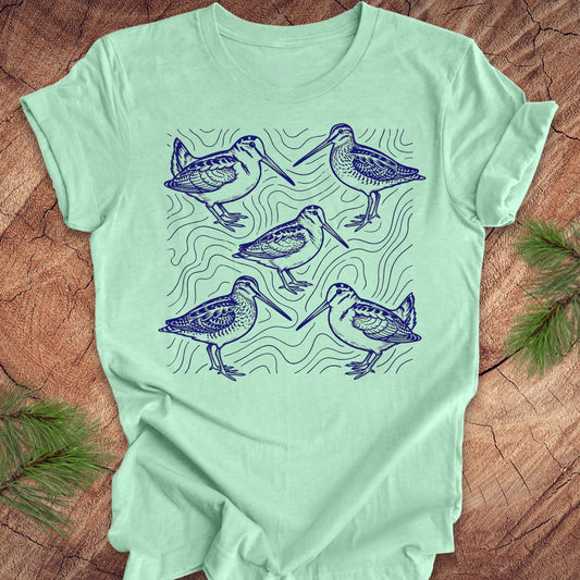 Mint green t-shirt with blue woodcock and wilson's snipe bird illustrations on a wooden surface