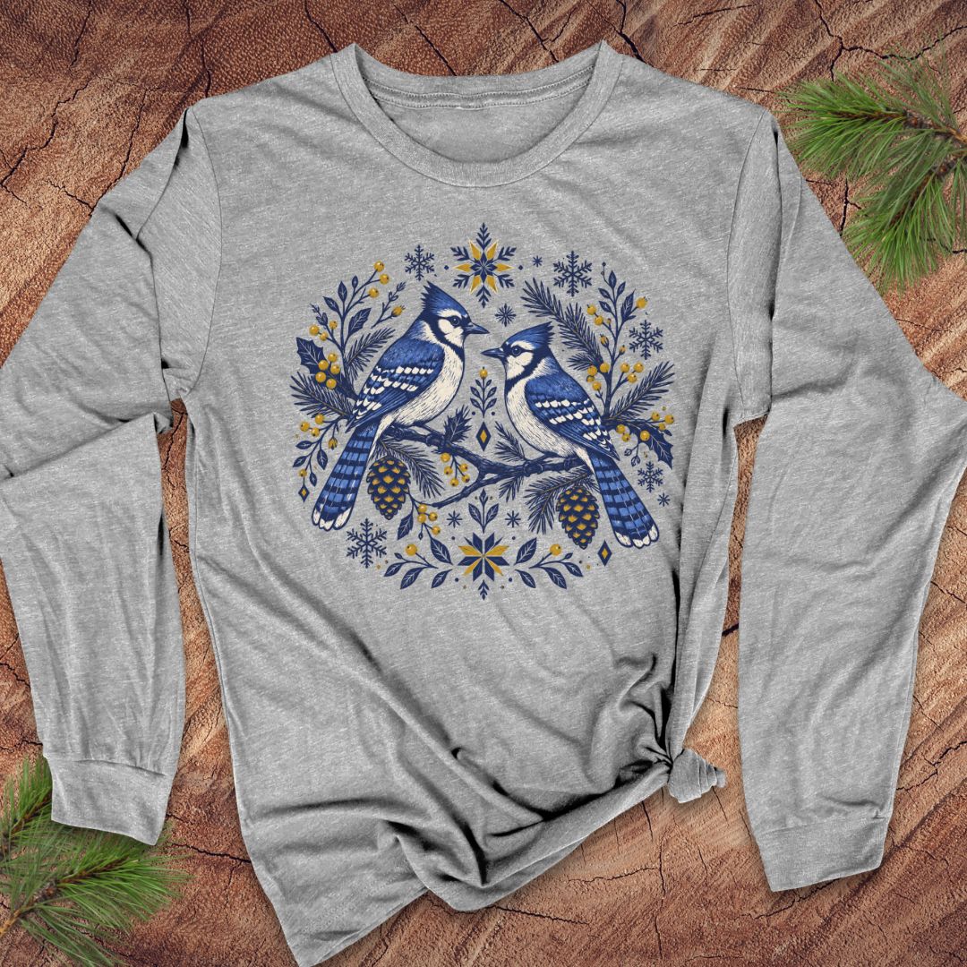 A heather gray long-sleeve t-shirt with a blue jay graphic, laid flat on a rustic wood background with pine needles.