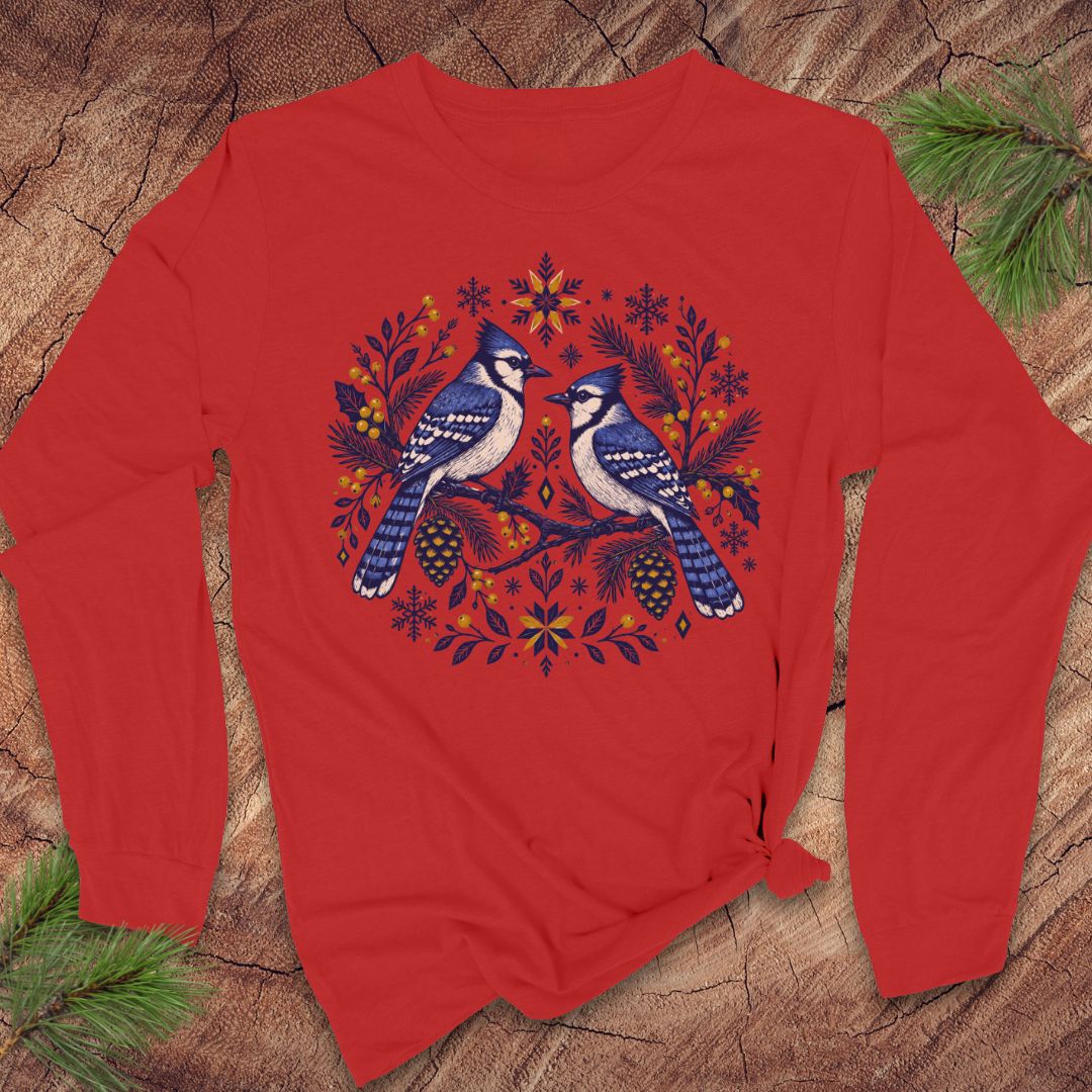A red long-sleeve crewneck sweatshirt featuring a graphic of two blue jays surrounded by festive autumn and winter motifs like pinecones, snowflakes, and evergreen branches, laid flat on a rustic wood background with pine needles.