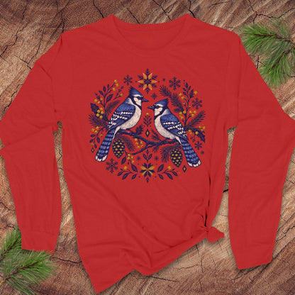 A red long-sleeve crewneck sweatshirt featuring a graphic of two blue jays surrounded by festive autumn and winter motifs like pinecones, snowflakes, and evergreen branches, laid flat on a rustic wood background with pine needles.
