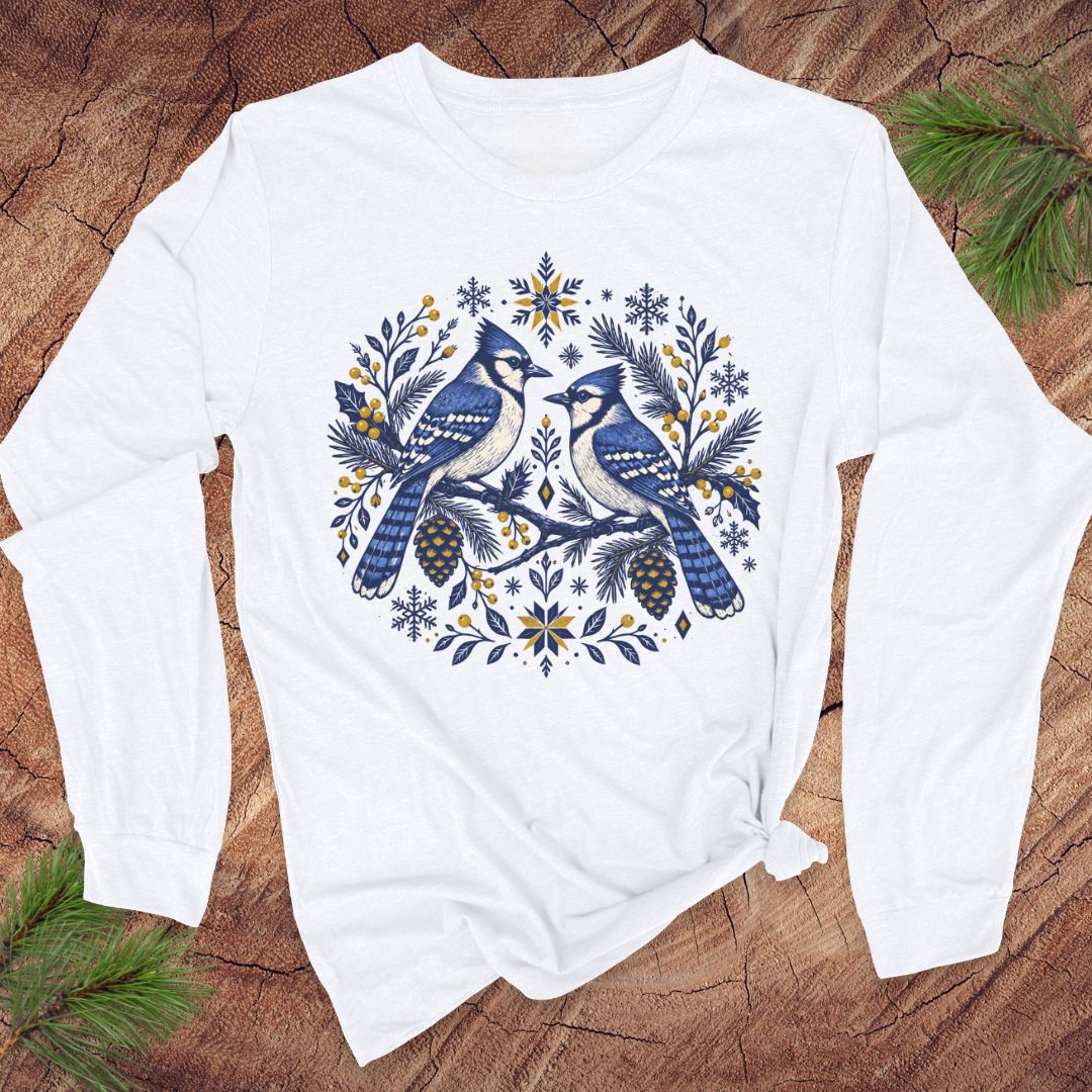 A white long-sleeve t-shirt with a circular graphic of two blue jays surrounded by pinecones, pine needles, and snowflakes, laid flat on a rustic wood background with pine branches.