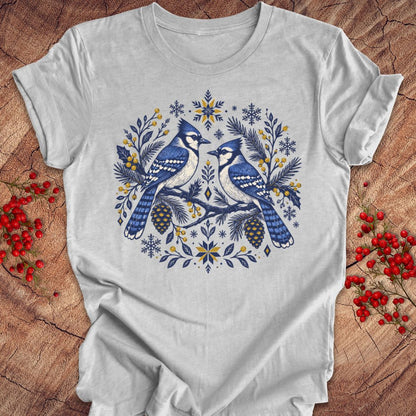 A light gray short-sleeve crewneck t-shirt featuring a detailed graphic of two blue jays surrounded by pinecones, pine needles, and snowflakes in blue, white, and yellow. The t-shirt is laid flat on a rustic wooden surface decorated with red berries.