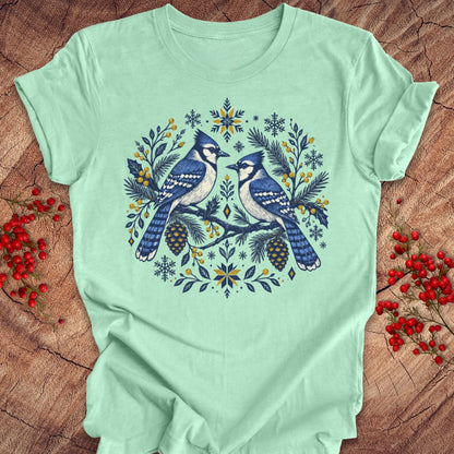 A mint green short-sleeve t-shirt with a detailed graphic of two bluebirds in a circular wreath design with pinecones, snowflakes, and floral elements, laid flat on a rustic wood background with red berries.